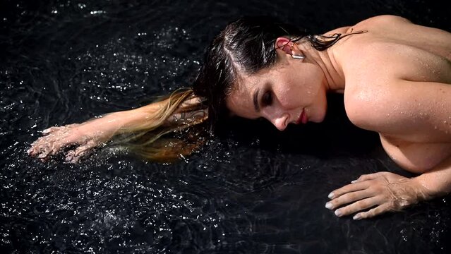 Seductive pretty wet young lady squeezes wet clothes, top standing under rain shower slow motion isolated on black studio background. Aqua studio 4k video slow motion 120 fps raw footage