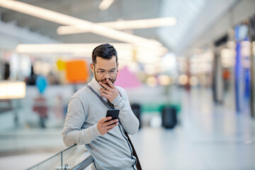 A young man is standing at the shopping mall and carefully reading messages on the phone.