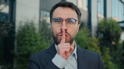 City portrait Caucasian business man businessman outdoors showing silence hush lips gesture keep secret secrecy confidential information stop talk silent quiet put finger to mouth mysterious hand sign - obrazy, fototapety, plakaty