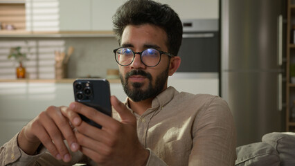 Arabian Indian ethnic man guy use mobile phone on couch browsing mobile internet at home kitchen...