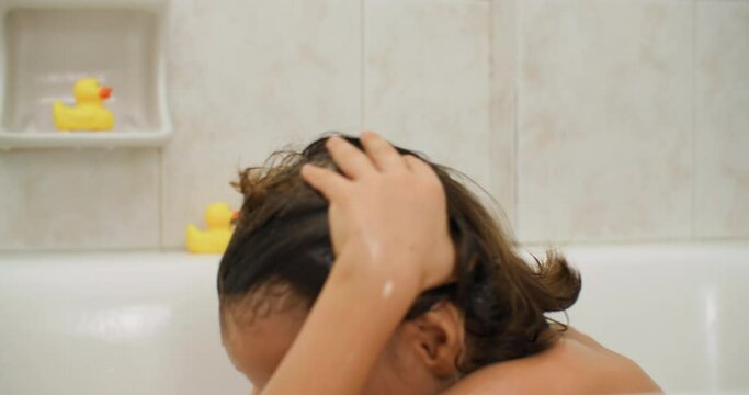 A girl child with wet hair is bathing in the bathroom. Happy smiling child having fun and fooling around with water in the bathroom. High quality 4k footage