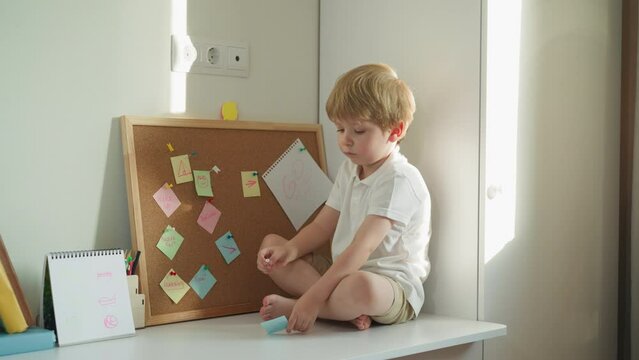 Cute blond boy sits on desk near cork board and window. Barefoot toddler examines piece of paper with note holding pin. Preschooler rests in home study room