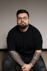 Man in a black t-shirt and ripped jeans, sitting on a stool. He has tattoos on both arms. The...