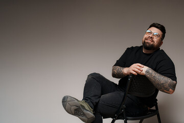 A photo of a tattooed man sitting on a black chair with his legs crossed. He is wearing a black t-shirt and black jeans. This image can be used for advertising, editorial, or personal projects. - Powered by Adobe