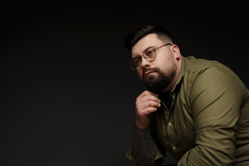 Obraz premium A portrait of a thoughtful bearded man in glasses wearing a green shirt. He is resting his chin on his hand and looking away from the camera. The background is dark gray