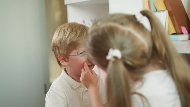 Boy laughs while girl paints face slow motion. Sister draws glasses and lines around nose. Friendly couple of kids plays with body painting closeup