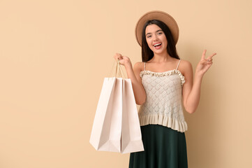 Happy beautiful young woman with shopping bags showing victory gesture on beige background