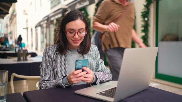 Woman in jacket and horn-rimmed glasses typing a message on mobile phone. Attractive young female using cell phone and notebook sitting in outdoor cafe backdrop store windows decorated for Christmas