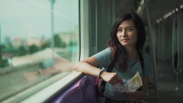 Cinematic Close Up Shot Of Young Woman Looking Through Window While Traveling By Train Through City Landscape. Map In Hands. Nostalgic Female Feeling Homesick And Smiling While Reaching Her Station