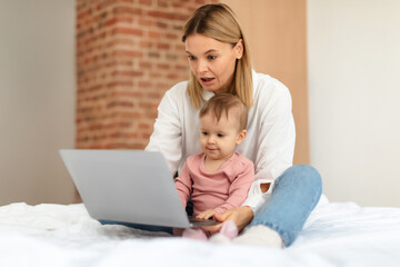 Baby and gadgets. Mommy and her little daughter using laptop, browsing internet for online fun, sitting on bed at home