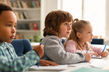 Elementary classroom of diverse children listening attentively to their teacher, writing in...