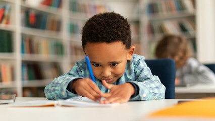 Concentrated black schoolboy sitting at desk in school and learning to write in copybook with...