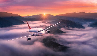 Airplane is flying above mountain peak in pink low clouds at sunrise. Landscape with passenger...