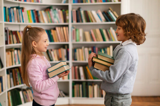 School Boy And Girl Holding Stack Of Books And Talking With Each Other Standing In Library, Side View