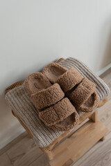 Fototapeta premium Stack of warm neutral beige clothes on wooden stool over white wall. Fluffy slippers, wool pullovers and sweaters. Minimalist autumn, fall fashion background