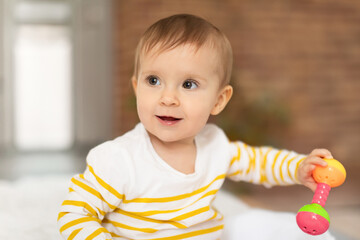 Infant baby girl playing with rattle toy while sitting on bed at home, looking aside and smiling