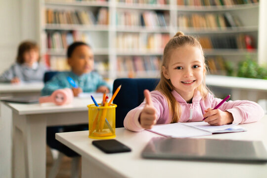 Smart girl doing thumbs up gesture while studying and sitting at desk in classroom, ready to study for excellence