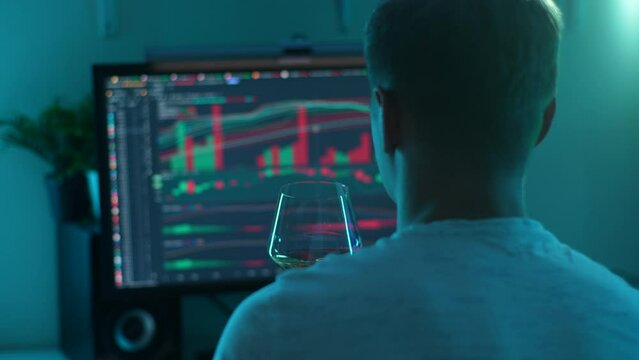 Back view of unrecognizable successful male stock trader celebrating drinking wine watching real-time stocks, exchange market charts on desktop monitor. Man works in investment at home office.