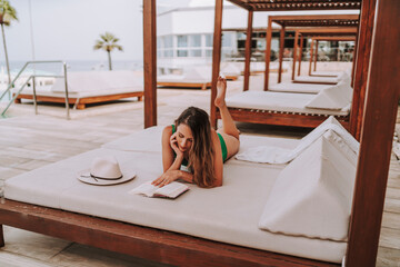 Chica joven delgada en cama balinesa descansando y leyendo tranquilamente  - obrazy, fototapety, plakaty