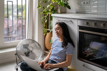 Relaxed Asian woman typing on laptop cooling by floor electric fan sits lotus position on floor in kitchen. Carefree Chinese girl blogger inspiredly writes article online magazine, work at home