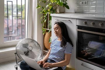 Tuinposter Relaxed Asian woman typing on laptop cooling by floor electric fan sits lotus position on floor in kitchen. Carefree Chinese girl blogger inspiredly writes article online magazine, work at home © DimaBerlin