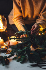 Young woman in knitted sweater doing handmade Christmas wreath from natural materials: fir tree branches, dry citrus slices, cones, cinnamon sticks. Wooden table, cozy light, home atmosphere