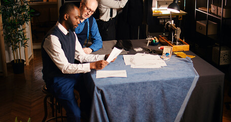 Designer measuring customers body, using industrial meter to ensure right size for custom made order. Senior tailor in atelier discussing with client about suit design. Handheld shot.