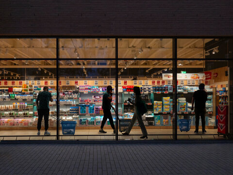 A look through a window in a supermarket at night time with people with people walking around shopping, looking at goods and talking on their mobile phones