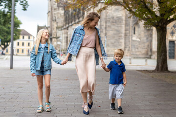 Fototapeta na wymiar Young adult beautiful mom enjoy having fun walking with two kids in european city street at summer day. Happy parenthood family concept. Mother care resonsibility. People go outside in Germany