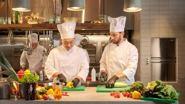 Two multicultural chefs communicate at workplace on background of restaurant professional kitchen. Asian and european cooks cutting vegetables and prepearing vegetarian dishes.