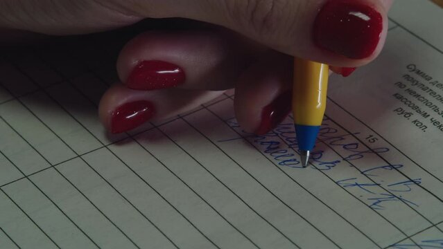 Close-up of female hands with red painted nails that quickly cursive entries with a yellow pen with blue ink in a ledger. Working in an office where a left-handed girl writes from left to right