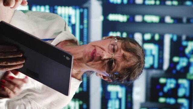 Vertical video Organized admins in high tech workspace, completing tasks on tablet while running scripts on laptop terminal. Focused specialists amidst server clusters, accessing databases and