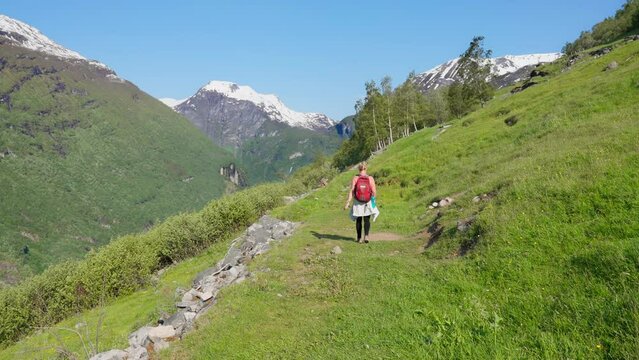 Following behind a woman looking out on the mountain peak fjords and valley below in a grassy meadow in Geiranger, Norway