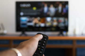 Hand holding remote control while searching for a series on television - obrazy, fototapety, plakaty