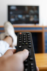 Vertical photo of a hand holding a television remote control while his feet are on the table.