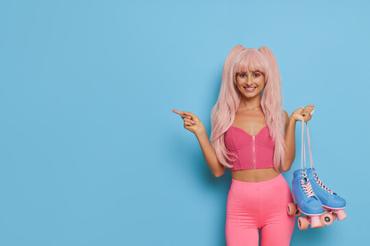 Smiling girl in pink clothes and pink hair holding blue roller-skates and points aside, picture setup on blue background, vintage concept, copy space