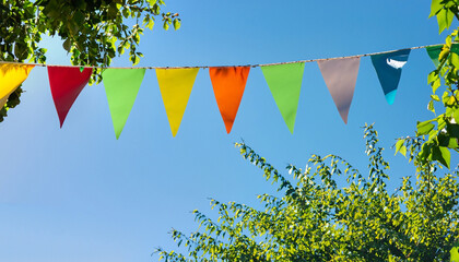 colorful pennant string decoration in green tree foliage on blue sky, summer party background template banner with copy space