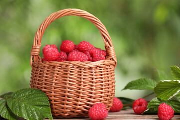 Fototapeta na wymiar Wicker basket with tasty ripe raspberries and leaves on wooden table against blurred green background, closeup
