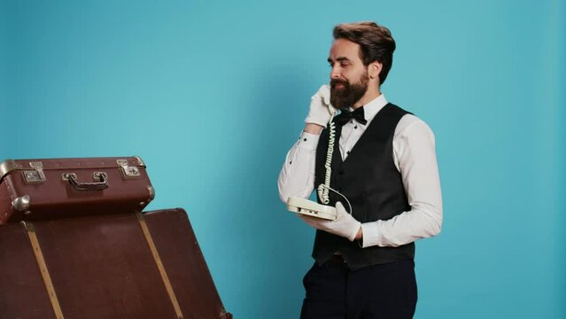 Classy bellhop using landline phone on camera, talking to guests about booking accommodation. Skilled professional porter making reservations remotely with office cord telephone.