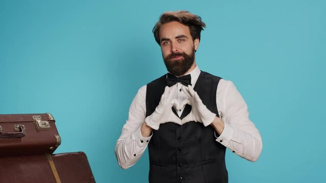 Cheerful bellhop does heart shaped sign in studio, expressing romantic honest feelings and flirting while he wears formal suit. Lovely employee showing romance symbol for love, sincere emotions.