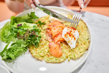 Woman cuts Zucchini pancakes, with avocado, cheese cream, salmon and egg . Healthy breakfast, protein. Restaurant dish. Breakfast in cafe on a sunny morning, on the summer veranda