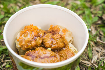 Indonesian Siomay or Dim Sum - Steamed shrimp shumai dumpling dimsum with peanut and chilli sauce in white paper bowl on lawn grass background.