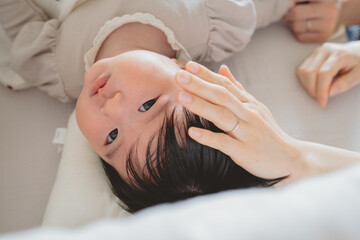 Fototapeta na wymiar 春の日中、ベッドに横になり母親に頭を撫でられるアジア人の黒髪の赤ちゃん
