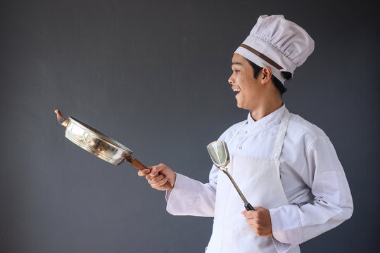 Male chef in white uniform and hat holding frying pan as juggling food during cooking process on grey background