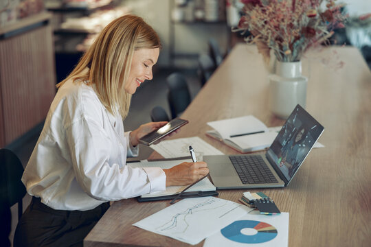 Smiling business woman working on laptop, making notes in cozy coworkign space interior