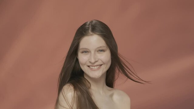 A seminude woman demonstrates healthy silky hair in a studio on a red background. The straight, long hair of a woman flutters in the wind. Keratin straightening. Slow motion. HDR BT2020 HLG Material.