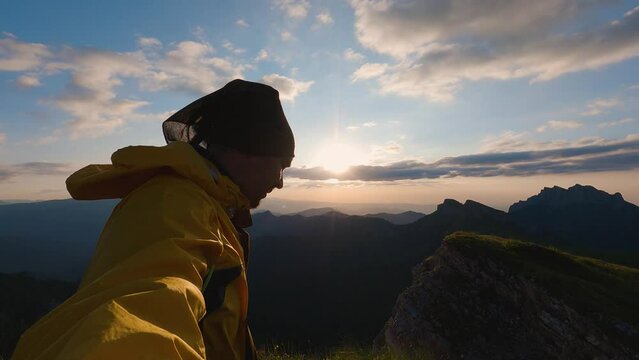 Tourist in yellow windbreaker and buff walking in the mountains with sunset in the background. A man looks into the camera and talks about walk. Beautiful sunset sky and mountain peaks in distance