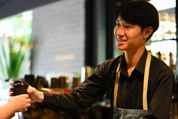 Fototapeta na wymiar Smiling man barista wearing apron standing behind counter and serving coffee to customer. Start up, small business owner concept