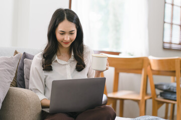 A millennial Asian businesswoman is sitting and working with laptop computer in her home kitchen, while also enjoying a relaxing cup of coffee.