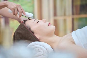 아시아 한국의 젊은 여성이 스파 또는 피부관리전문점 또는 피부과 병원에서 침대에 누워 눈을 감고 고주파마사지 미용기계로 전문관리사에게 얼굴부분에 리프팅 또는 주름관리를 받고 있다.